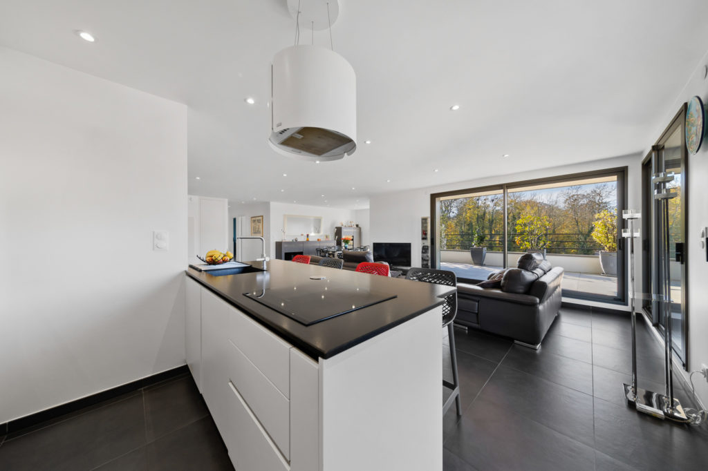 Cuisine moderne avec îlot central blanc et plan de travail noir, ouverte sur un salon lumineux et une terrasse.