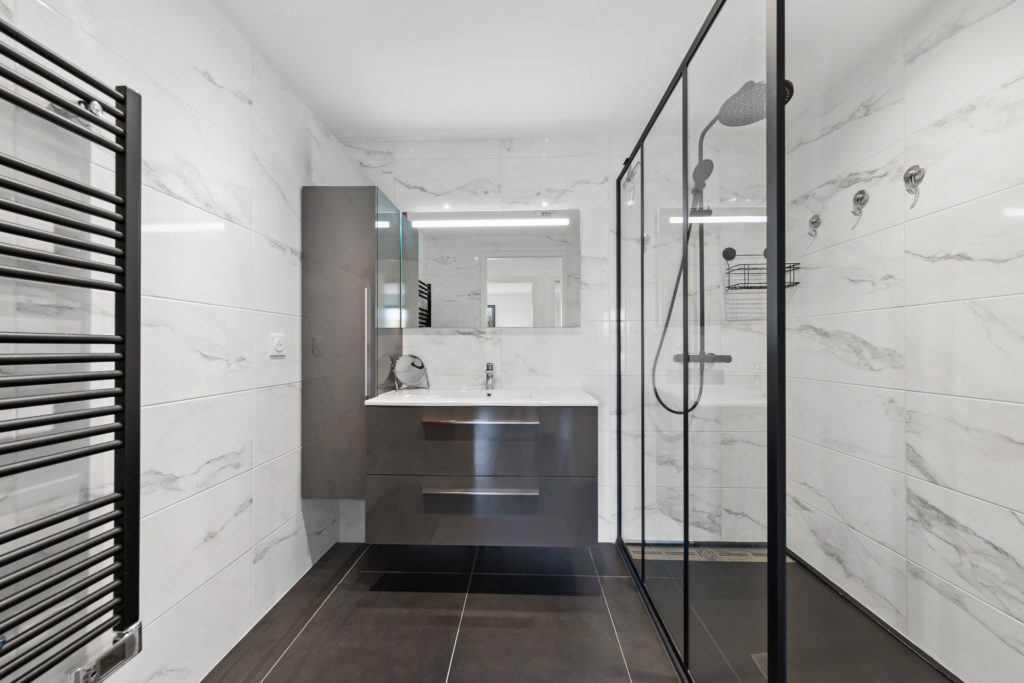 Salle de douche moderne avec miroir lumineux LED, robinetterie noire mate et revêtement mural en pierre naturelle.