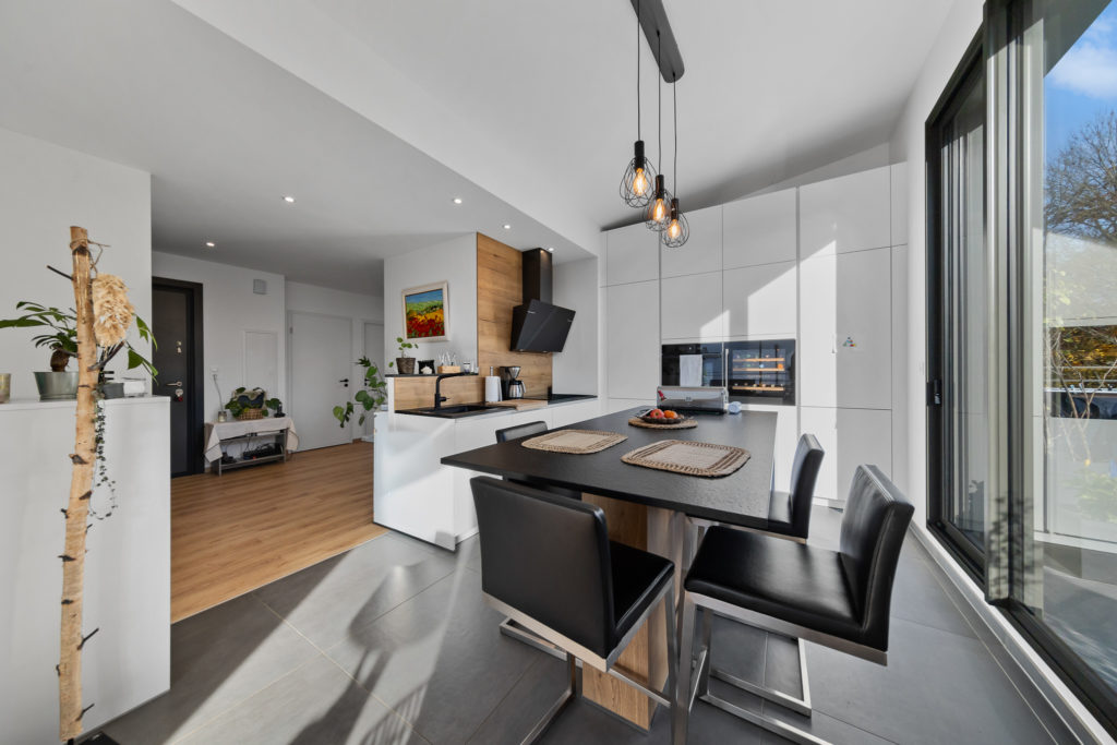 Cuisine haut de gamme avec coin repas, mobilier intégré blanc brillant et crédence en bois, baignée de lumière naturelle.